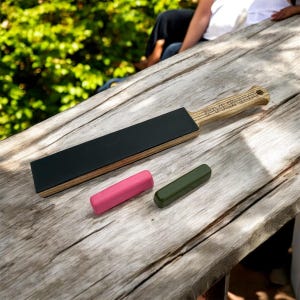 Può includere: Una strop in legno con una superficie in pelle nera e un manico in legno con la parola "PHURFER" incisa su di esso. Sono anche mostrati due piccoli blocchi di cera colorata, uno rosa e uno verde.