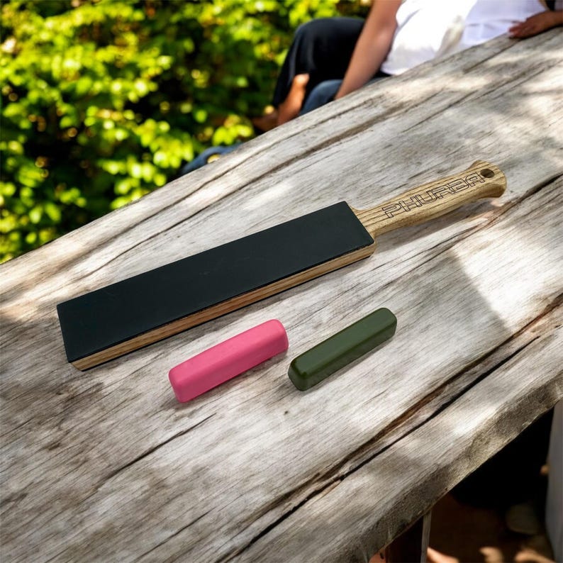May include: A wooden strop with a black leather surface and a light-colored handle with the word "PHURBA" engraved. Two small, cylindrical sharpening compounds, one pink and one olive green, are also present.