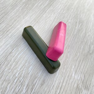 May include: Two rectangular wax blocks. One is a dark olive green, and the other is a bright pink. The pink block rests on top of the green block, creating a V-shape. The blocks are on a light wood-grain surface.