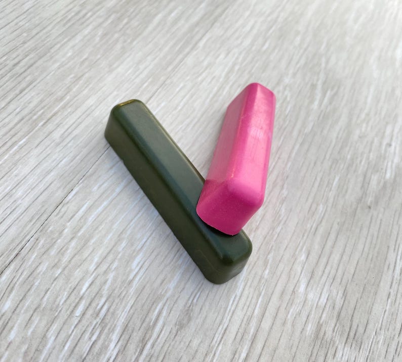 May include: Two rectangular wax blocks. One is a dark olive green, and the other is a bright pink. The pink block rests on top of the green block, creating a V-shape. The blocks are on a light wood-grain surface.