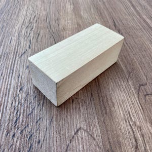 以下が含まれることがあります： 滑らかな表面を持つ、明るい色の長方形の木製ブロック。ブロックは、濃い茶色の木目調の表面に斜めに配置されています。木製ブロックは、より大きなアイテムのコンポーネントのようです。