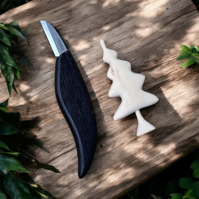 以下が含まれることがあります： 黒い木製の柄と銀色の刃が付いた彫刻刀と、小さな明るい色の木製ツリーの置物。 ナイフと木は木製の表面にあります。