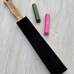 May include: A wooden strop with a black sharpening surface and the word "PHURBA" engraved on the handle. Two sharpening compounds, one green and one pink, are also present. The items are arranged on a light wood grain surface.
