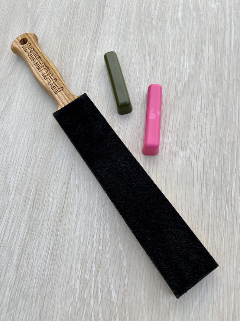 May include: A wooden strop with a black sharpening surface and the word "PHURBA" engraved on the handle. Two sharpening compounds, one green and one pink, are also present. The items are arranged on a light wood grain surface.