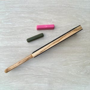 May include: A wooden fan with a light brown handle and a black and light brown fan blade. Two rectangular objects, one pink and one green, are also in the image. The fan is angled diagonally across a light gray wooden surface.