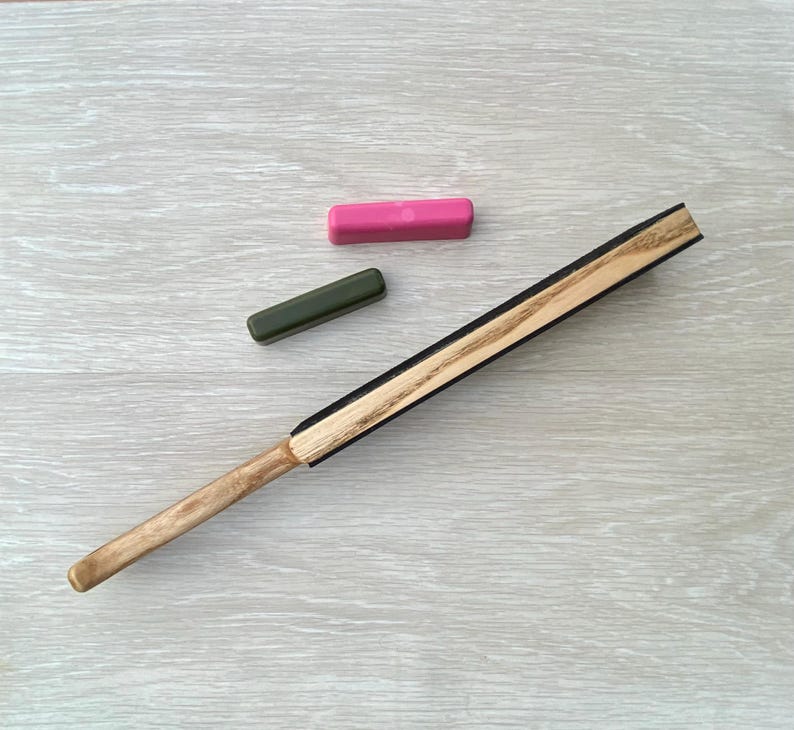 May include: A wooden fan with a light brown handle and a black and light brown fan blade. Two rectangular objects, one pink and one green, are also in the image. The fan is angled diagonally across a light gray wooden surface.