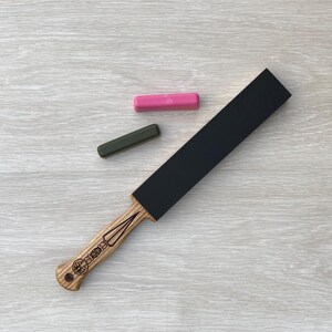 May include: A straight razor strop with a black leather surface and a wooden handle. The handle has a decorative design and is made of light-colored wood. Two sharpening compounds, one pink and one green, are also present.
