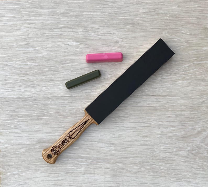 May include: A straight razor strop with a black leather surface and a wooden handle. The handle has a decorative design and is made of light-colored wood. Two sharpening compounds, one pink and one green, are also present.