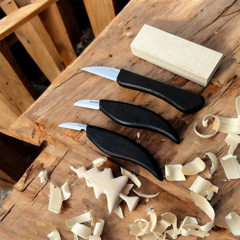 以下が含まれることがあります： 黒い柄と銀色の刃を持つ木彫り用のナイフが3本、木製の表面に配置されています。木片と木くずも一緒に写っています。木彫りのクリスマスツリーもあり、クラフトのテーマを示唆しています。