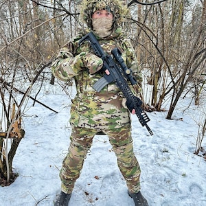 May include: A person wearing camouflage clothing and a ghillie suit holds a rifle in a snowy forest setting.