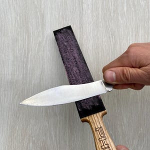 May include: A knife being sharpened on a purple strop. The knife has a silver blade and a wooden handle with the word "PHUR" engraved on it. The strop is black and purple.