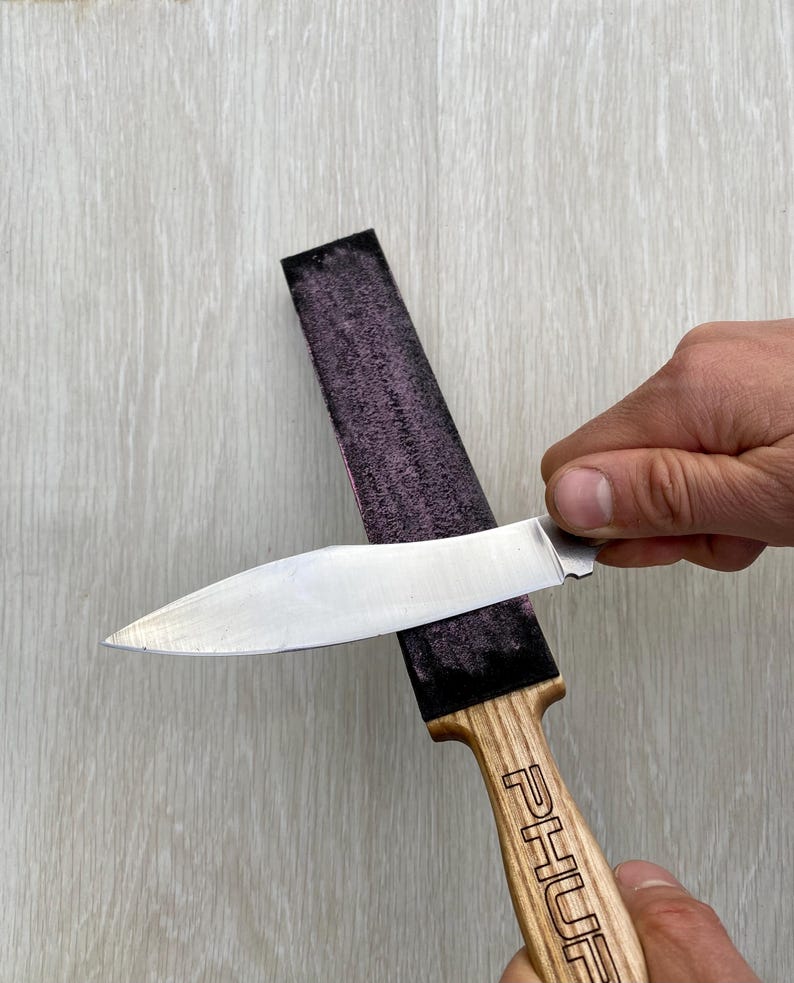 May include: A knife being sharpened on a purple strop. The knife has a silver blade and a wooden handle with the word "PHUR" engraved on it. The strop is black and purple.