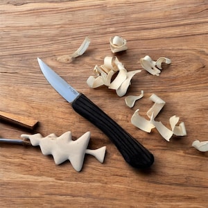 May include: A close-up of a wooden table with a small wooden Christmas tree, wood shavings, and a black handled knife with a silver blade. The knife has the word "F.Dick" printed on the blade.