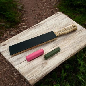 Puede incluir: Un asentador de madera con una superficie de cuero negro y un mango de color claro, junto con dos compuestos de afilado cilíndricos, uno rosa y otro verde oliva, sobre una superficie de madera. El mango tiene la palabra "PHURBA" grabada.