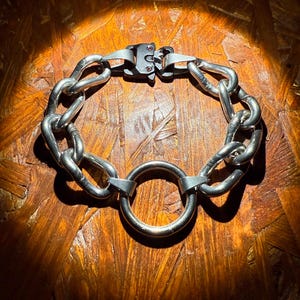 May include: A silver-toned chain bracelet with a large circular ring and a clasp closure. The bracelet features thick, rounded links and a prominent central ring. The clasp has small red accents. The jewelry is displayed on a wooden surface.