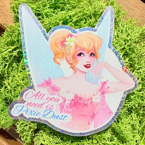 Puede incluir: Una pegatina holográfica que muestra a un hada rubia con una coleta alta, vistiendo un vestido rosa con flores y alas translúcidas. El texto "All you need is Pixie Dust" está impreso en letra cursiva rosa y azul en la parte inferior izquierda.