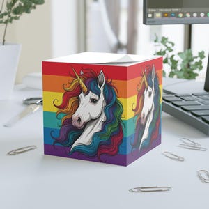 Può includere: Un blocco note color arcobaleno con un unicorno bianco con criniera e coda arcobaleno. L'unicorno guarda a destra. Il blocco note è su una superficie bianca.