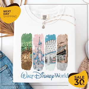 a white t-shirt with a colorful graphic design depicting various Disney landmarks, including the Walt Disney World resort, the Eiffel Tower, and the Hollywood Tower. The t-shirt is displayed on a white background, and there are yellow and black sale tags visible, indicating a 30% discount on the item.