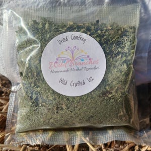 May include: A clear plastic bag filled with dried green herbs. A white circular label reads "Dried Comfrey" and "Wild Branches Homemade Herbal Remedies" with "Wild Crafted 1oz" below. The bag is resting on a bed of straw.