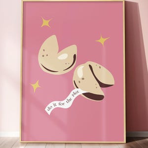 May include: A framed wall art print featuring a solid pink background with two beige fortune cookies. One cookie has a white paper strip extending from it with the black text "do it for the plot" printed on it. Three yellow four-pointed stars.