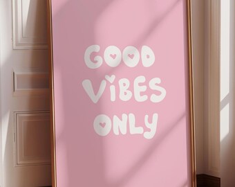May include: A framed pink art print with the words "GOOD VIBES ONLY" in white, whimsical lettering. The print has a gold frame and is leaning against a white wall and wooden floor. The artwork is a decorative piece.