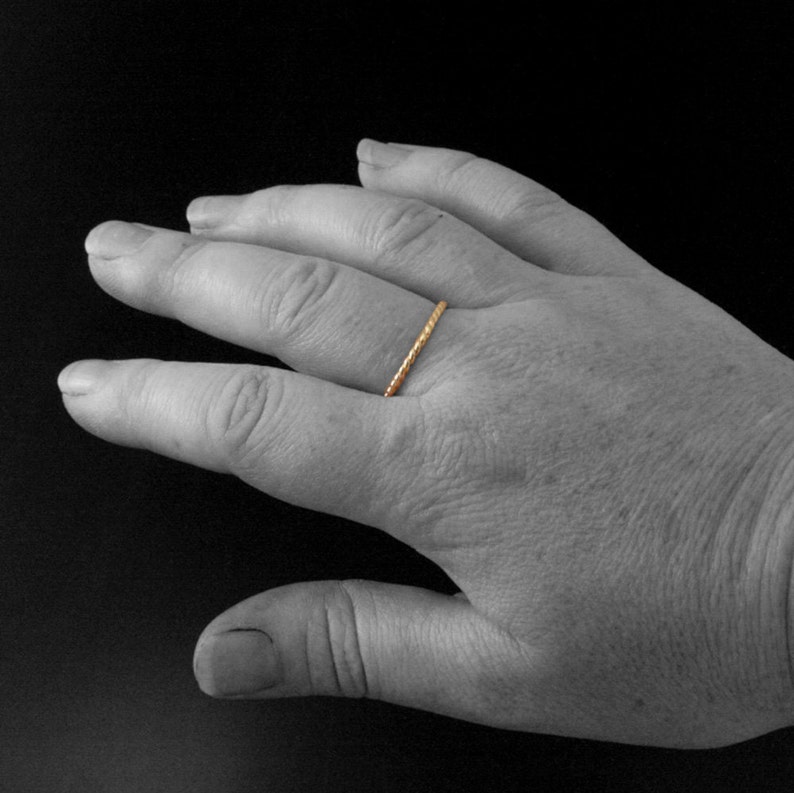 May include: A thin gold band ring with a textured surface. The ring is worn on the ring finger of a hand.