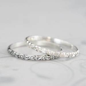 May include: Three silver rings with an ornate, swirling pattern. The rings are stacked, with the details of the design clearly visible. The rings are likely for a wedding or engagement.