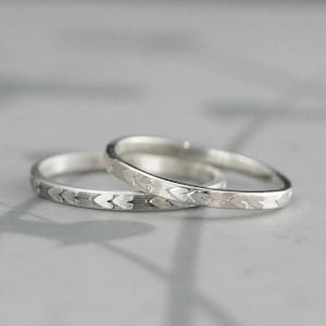 May include: Two silver rings with a repeating heart and line pattern. The rings are thin and sit on a light gray surface. The rings are likely wedding bands or promise rings.