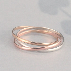 May include: Three intertwined rings in gold, rose gold, and silver tones. The rings are smooth and polished, creating a seamless, interlocking design. The jewelry is set against a soft, neutral background, highlighting the metallic colors.
