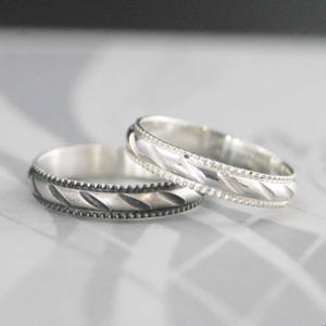 May include: Two silver rings with detailed patterns. One ring features a series of diagonal lines, while the other has a braided design. Both rings have a beaded border, creating a classic and elegant look.