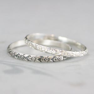 May include: Two silver rings with floral patterns. The rings are stacked on top of each other, with the top ring slightly out of focus. The rings have a detailed, engraved design of flowers and leaves.