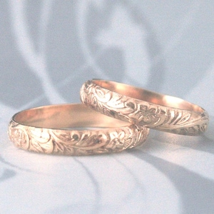Puede incluir: Dos anillos dorados con intrincados diseños florales y de hojas. Los anillos se exhiben sobre una superficie blanca y suave, mostrando sus grabados detallados y su cálido tono metálico. Ideales para una boda o compromiso.
