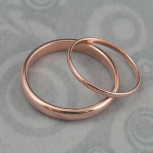 May include: Two simple rose gold wedding bands, one slightly larger than the other, are shown on a gray textured surface.