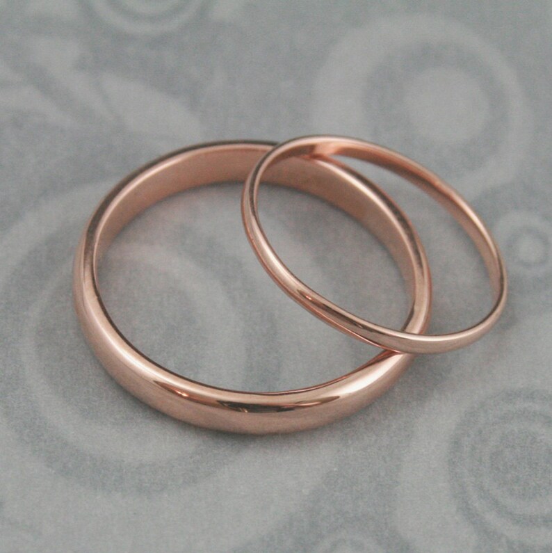 May include: Two simple rose gold wedding bands, one slightly larger than the other, are shown on a gray textured surface.