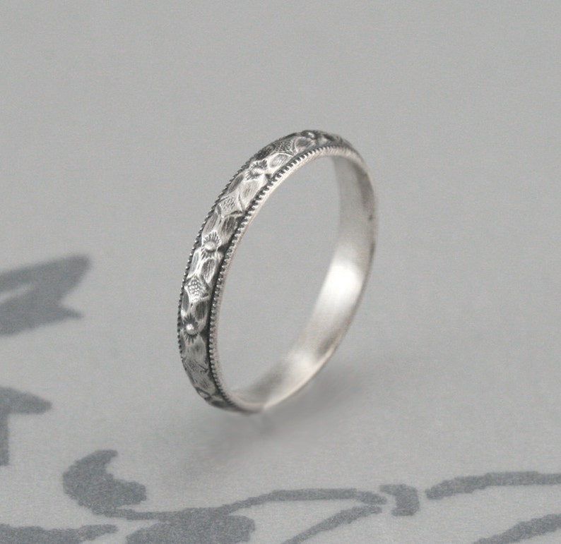 May include: A silver band ring with a delicate floral design engraved around the entire band.