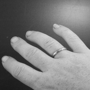 May include: A close-up of a hand wearing a thin silver band ring with a textured design.