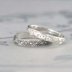 May include: Two silver rings with a repeating pattern of circles and floral designs. The rings are stacked on top of each other, with the top ring slightly angled. The rings are likely wedding bands or promise rings.