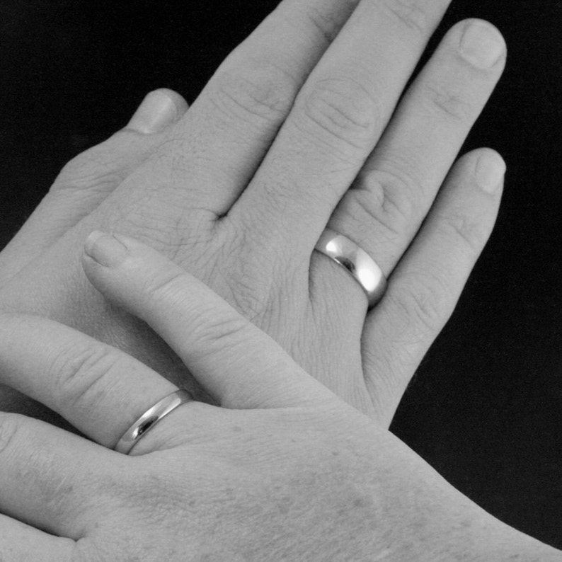 May include: Two hands with wedding bands on their ring fingers. The hands are clasped together, with one hand on top of the other.