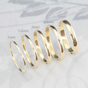 Può includere: Cinque anelli in oro lucido disposti in fila, ognuno con una larghezza di banda diversa etichettata come 1mm, 1,5mm, 2mm, 2,5mm e 3mm. Gli anelli hanno un profilo liscio e arrotondato con una finitura metallica riflettente, mostrando lo spessore.