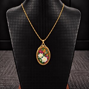 Peut inclure: Un collier pendentif ovale doré orné d'une illustration florale détaillée représentant des gardénias blancs et des oiseaux de paradis orange sur fond sombre, suspendu à une chaîne torsadée dorée assortie.