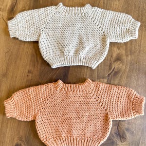 May include: Two handmade crochet sweaters. One is a light beige color, and the other is a soft peach. Both sweaters have a crew neck and long sleeves. The sweaters are displayed on a wooden surface.