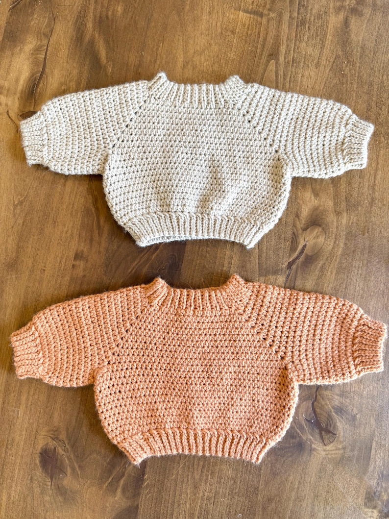 May include: Two handmade crochet sweaters. One is a light beige color, and the other is a soft peach. Both sweaters have a crew neck and long sleeves. The sweaters are displayed on a wooden surface.