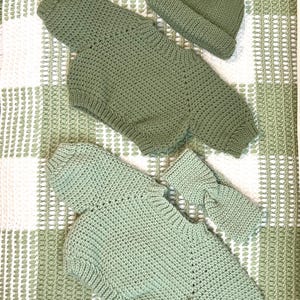 May include: Hand-crocheted baby outfit set in a soft sage green color. The set includes a romper with long sleeves and a matching beanie hat. The outfit is made of a textured yarn and is displayed on a white and green patterned surface.
