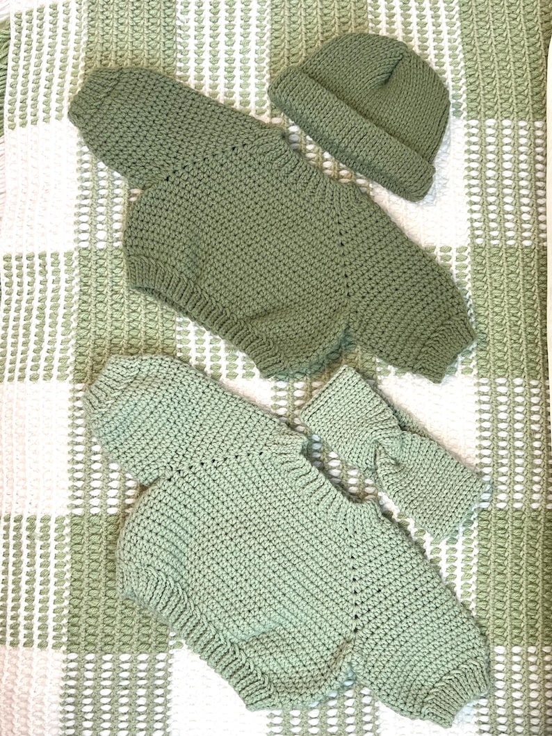May include: Hand-crocheted baby outfit set in a soft sage green color. The set includes a romper with long sleeves and a matching beanie hat. The outfit is made of a textured yarn and is displayed on a white and green patterned surface.