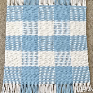 May include: A handmade, square blanket with a checkerboard pattern in light blue and white. The blanket has a fringe border in white and light blue. The blanket is made of a soft, textured material.