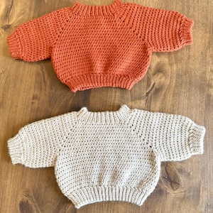 May include: Two handmade baby sweaters. One is a warm orange color, and the other is a neutral beige. Both sweaters have a round neckline and short sleeves. The sweaters are made of a textured, crocheted material.