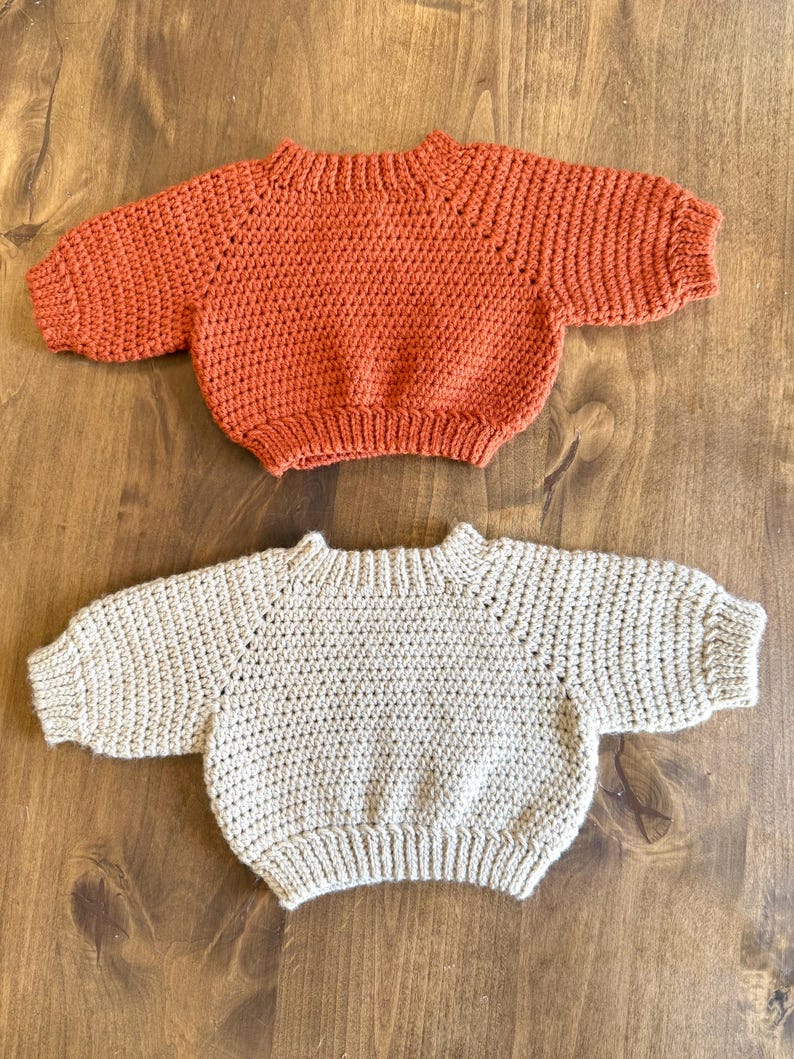 May include: Two handmade baby sweaters. One is a warm orange color, and the other is a neutral beige. Both sweaters have a round neckline and short sleeves. The sweaters are made of a textured, crocheted material.