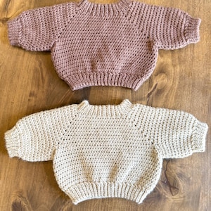 May include: Two baby sweaters. The top sweater is a dusty rose color, and the bottom sweater is a light beige. Both sweaters are crocheted with a round neckline and short sleeves. The sweaters are displayed on a wooden surface.
