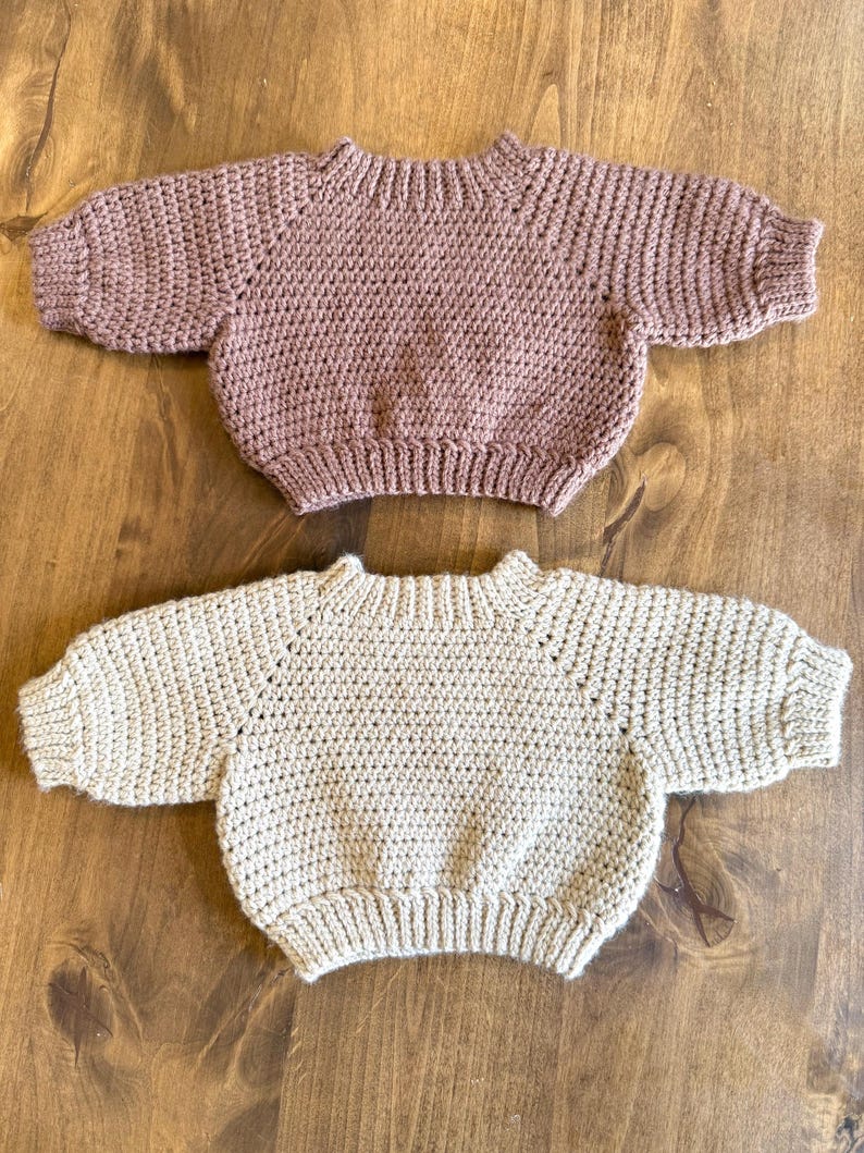 May include: Two baby sweaters. The top sweater is a dusty rose color, and the bottom sweater is a light beige. Both sweaters are crocheted with a round neckline and short sleeves. The sweaters are displayed on a wooden surface.