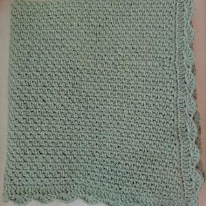 May include: A handmade, sage green crochet blanket. The blanket features a textured stitch pattern and a scalloped edge. The soft, muted green color and detailed craftsmanship suggest a cozy, comforting item.